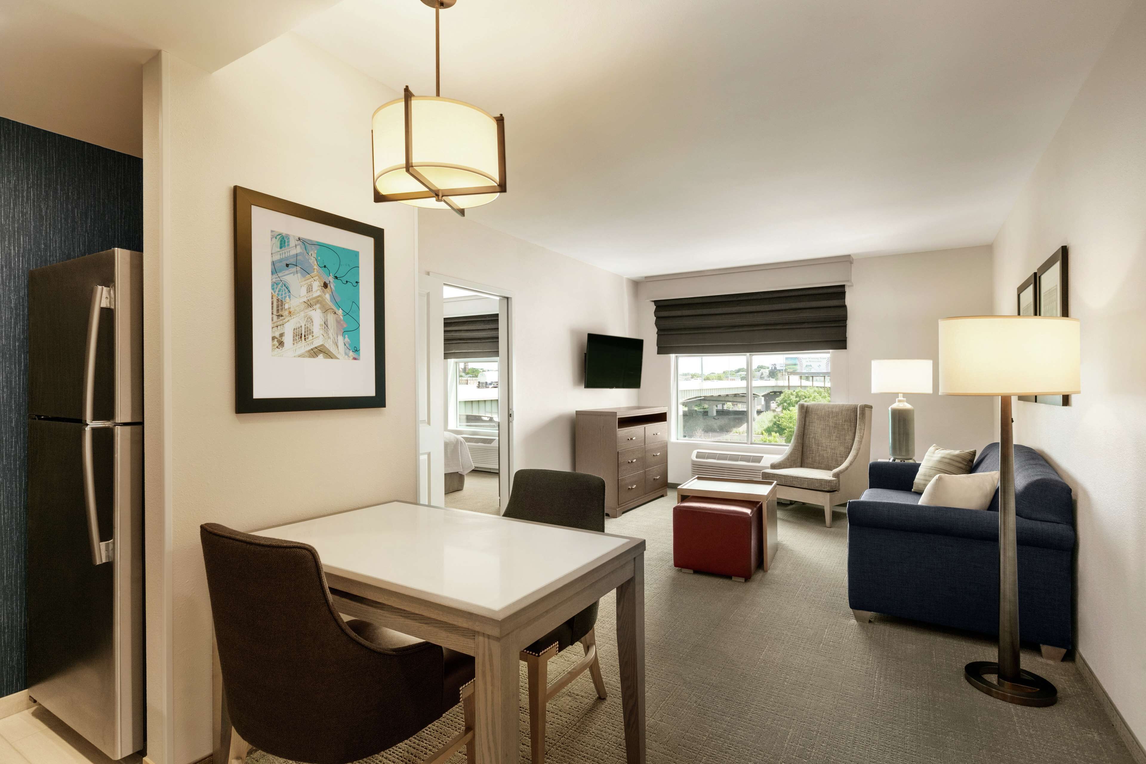 A modern hotel room with a dining area, a sofa, a TV, and a large window.