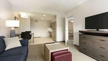 Una habitación de hotel moderna con sofá, televisión, zona de comedor y cocina al fondo.