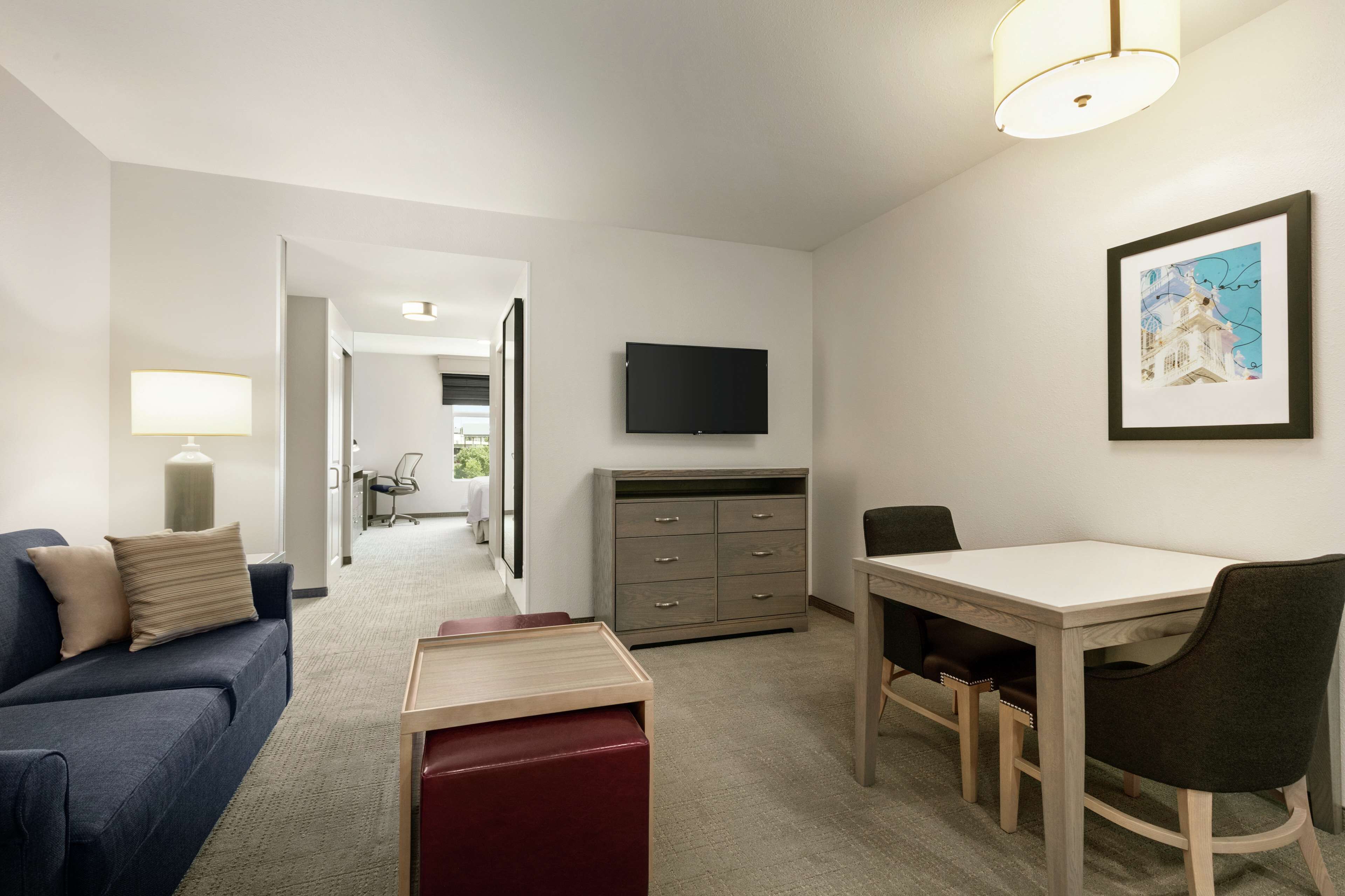 A hotel room with a sofa, a dining table with chairs, a TV mounted on the wall, and a desk with drawers.