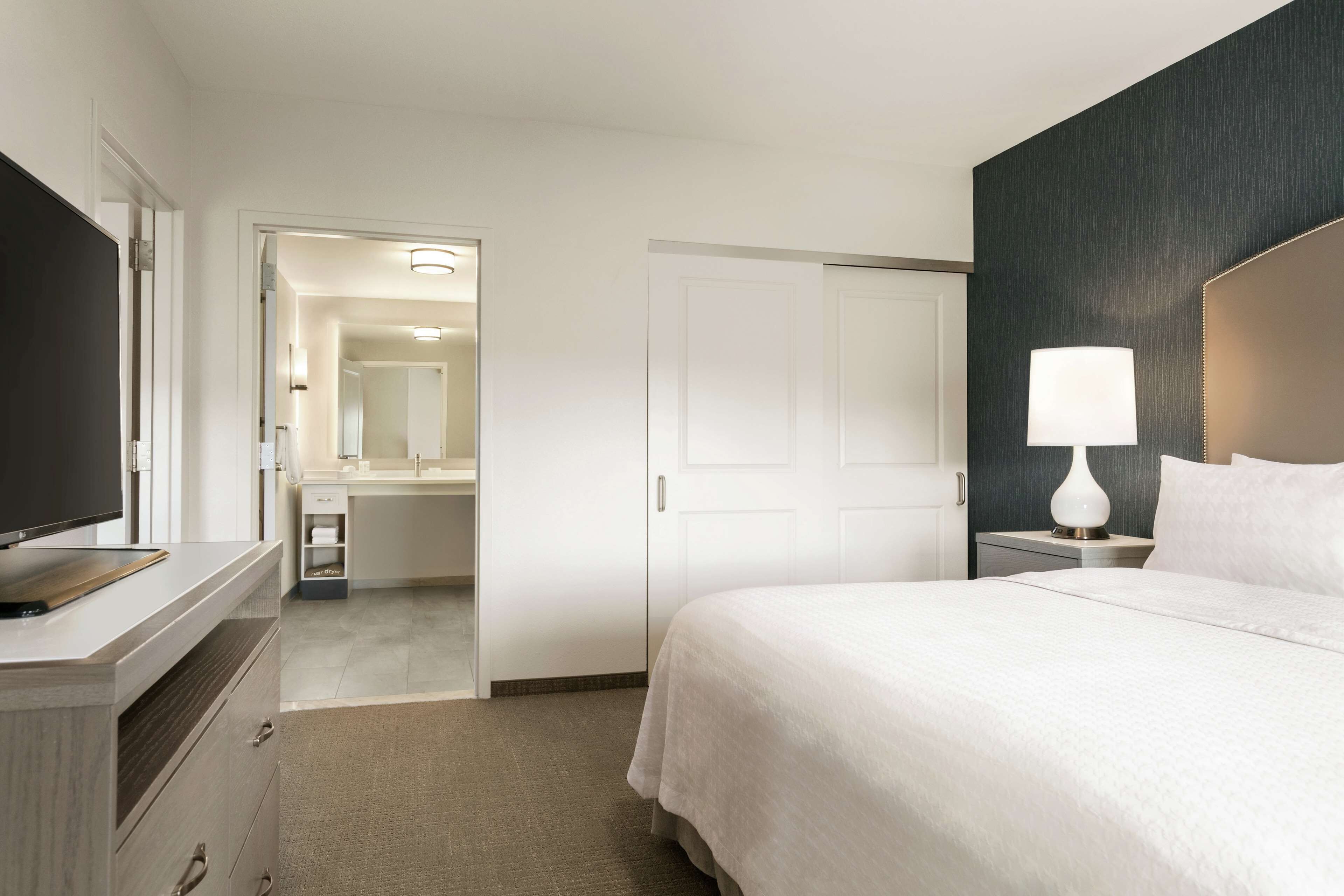 A hotel room with a bed, a TV, a desk, and a bathroom visible through an open door.