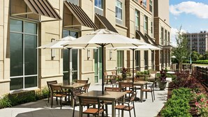 Een terras buiten met meerdere tafels en stoelen, omgeven door een gebouw met grote ramen en een bakstenen gevel.