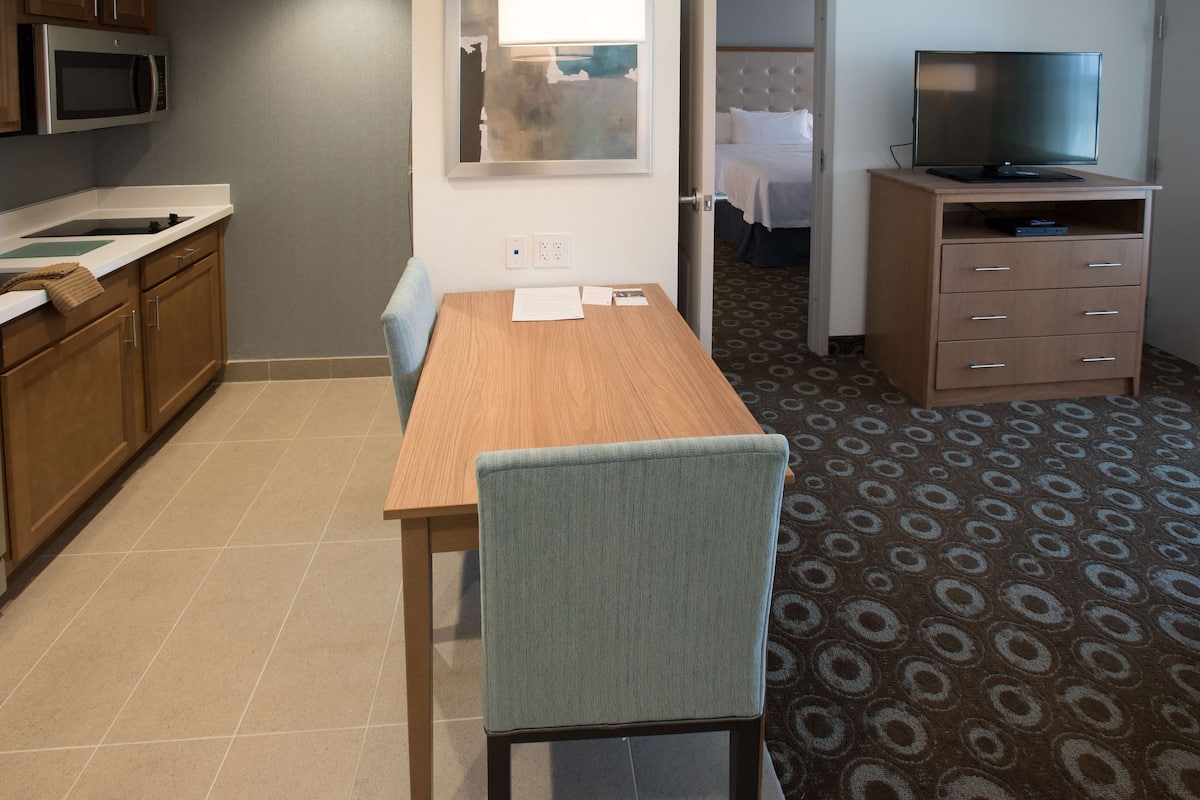 A hotel room with a wooden dining table, a blue chair, a TV on a wooden stand, and a patterned carpet.