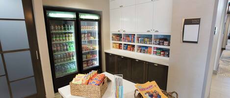 Un bar de aperitivos con un refrigerador lleno de bebidas y bocadillos, una exhibición de cajas de cereales y un cartel que anuncia los eventos del día.