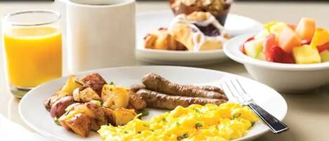 Ein Frühstücksteller mit Eiern, Würstchen und Kartoffeln, ein Glas Orangensaft, eine Tasse Kaffee, ein Croissant und eine Obstschale.