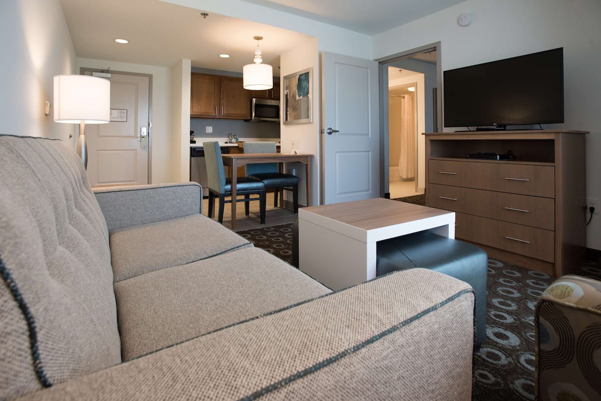 A living room with a sofa, a coffee table, a TV, and a kitchen area visible through an open door.
