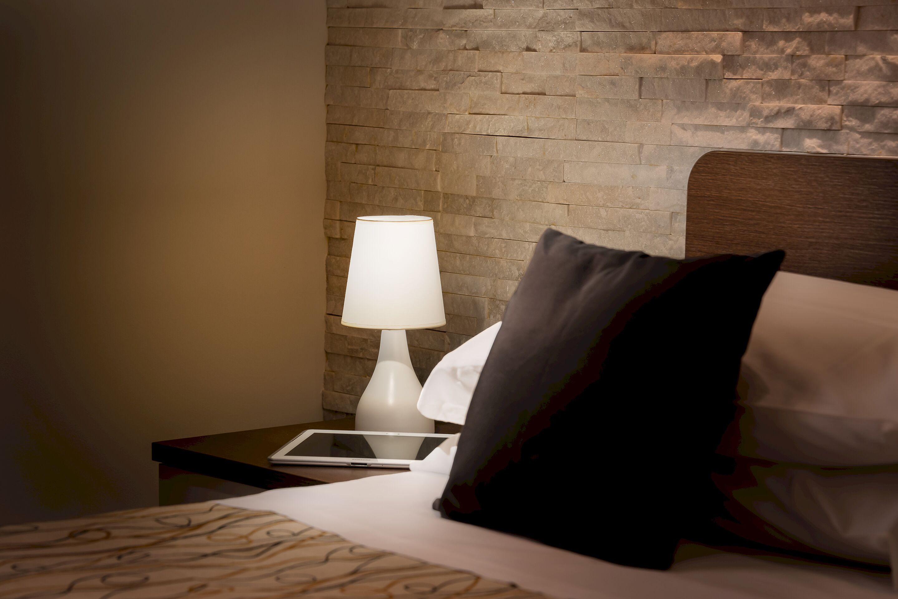 A neatly made bed with patterned bedding, a bedside lamp, a tablet, and a pillow with a dark and light color scheme.