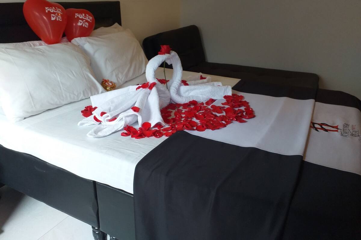 Uma cama com lençóis brancos, pétalas de rosa vermelhas e dois balões em formato de coração vermelhos, com a frase 'Feliz Dia' escrita neles.