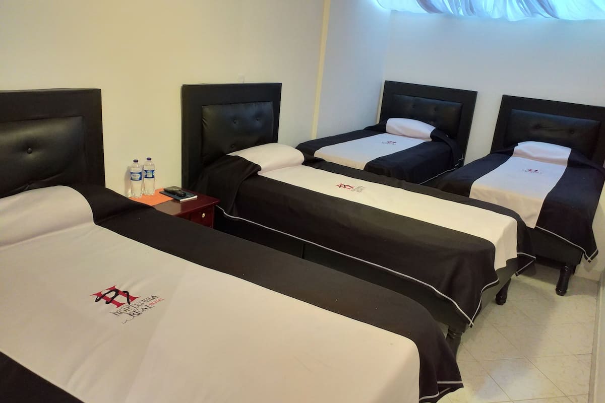 Quarto de hotel com três camas, cada uma com cabeceira preta e roupa de cama branca. Há uma pequena mesa com um abajur vermelho e uma garrafa de água.