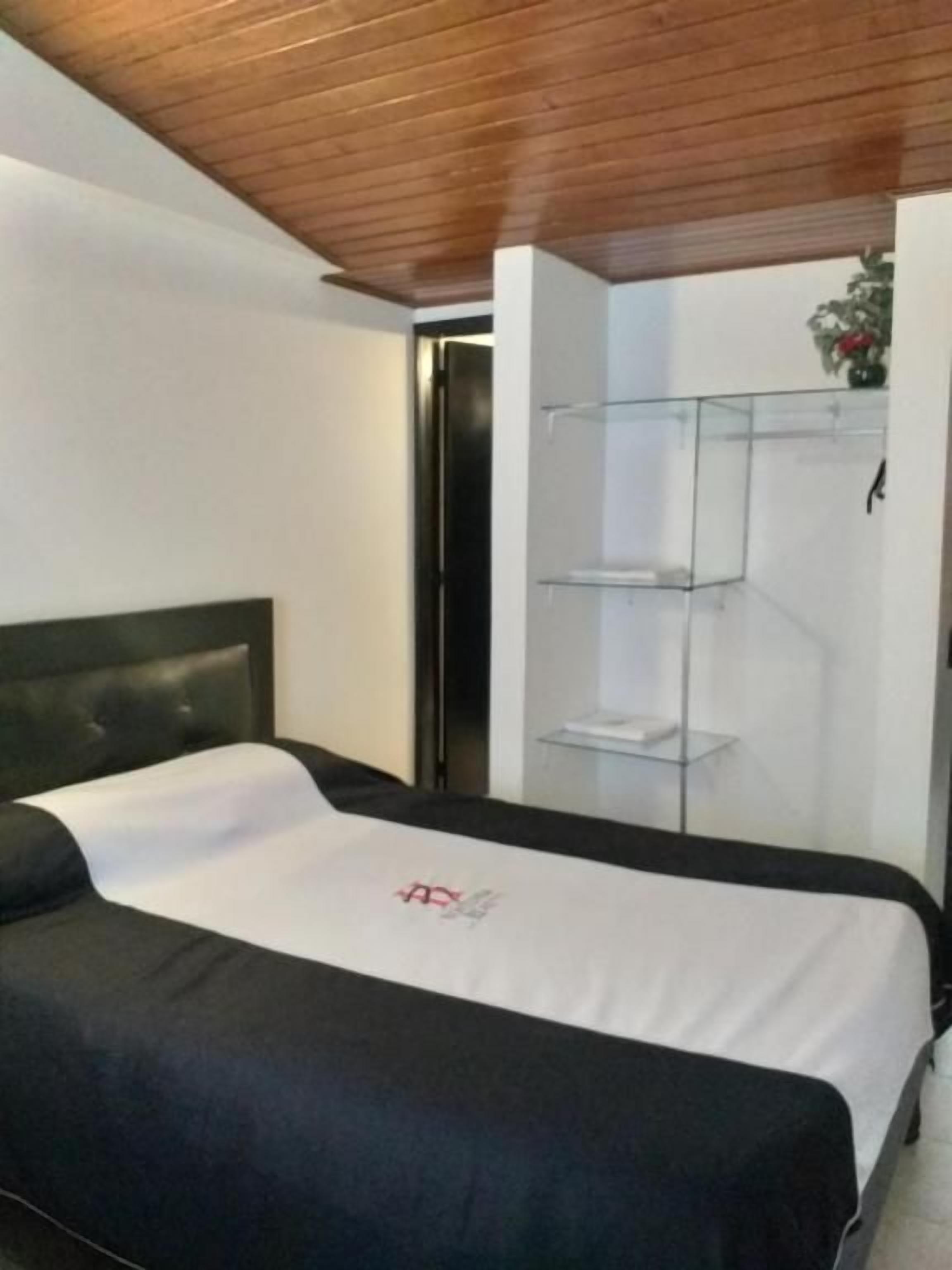 A bedroom with a bed, a wooden ceiling, a white wall, and a glass shelf unit.
