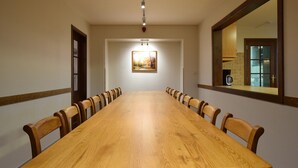 Una larga mesa de conferencias de madera con sillas, un cuadro en la pared y una cocina visible a través de una ventana.