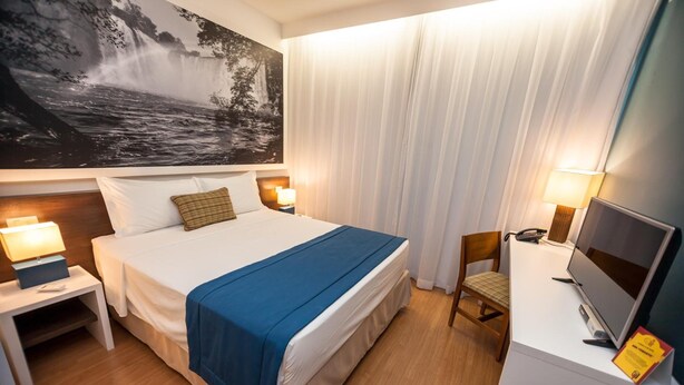 A hotel room with a large bed, two bedside tables with lamps, a desk with a computer and a TV, and a large painting of a waterfall on the wall. - Golden Tower Aeroporto - Lizz - By Fênix Hotéis (Uberlandia)