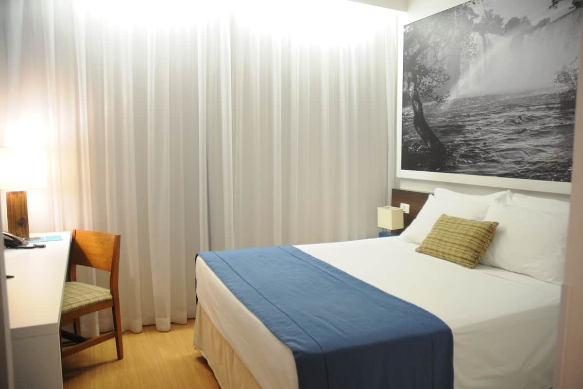 A hotel room with a bed, a desk, a chair, and a large framed picture on the wall.