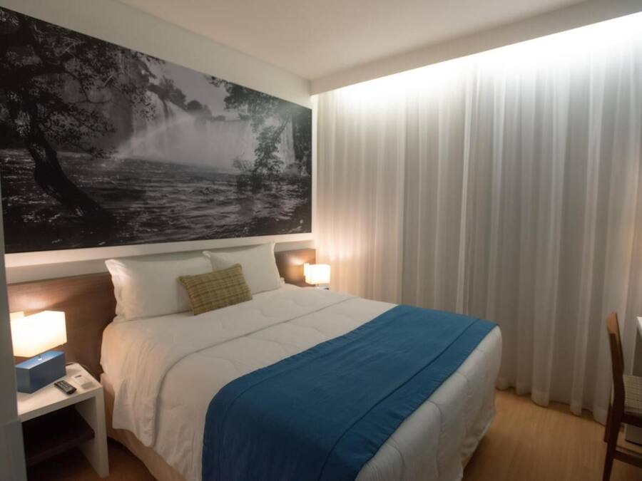 A hotel room with a large bed, two bedside lamps, a desk with a chair, and a large landscape painting on the wall.