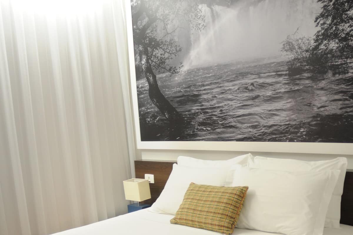 A hotel room with a bed, white bedding, a blue bedspread, a pillow, a nightstand with a lamp, and a large black and white waterfall painting on the wall.