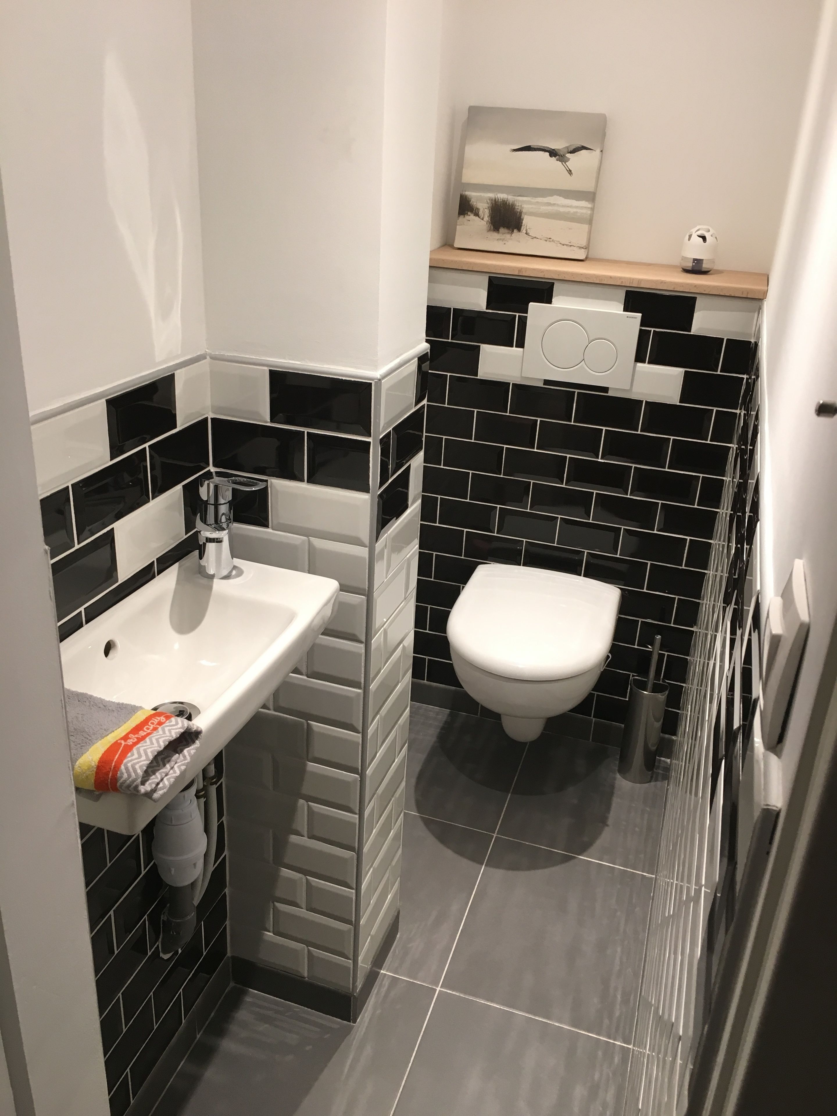 A modern bathroom with a white sink, black and white tiled walls, a toilet, and a small shelf with a picture.