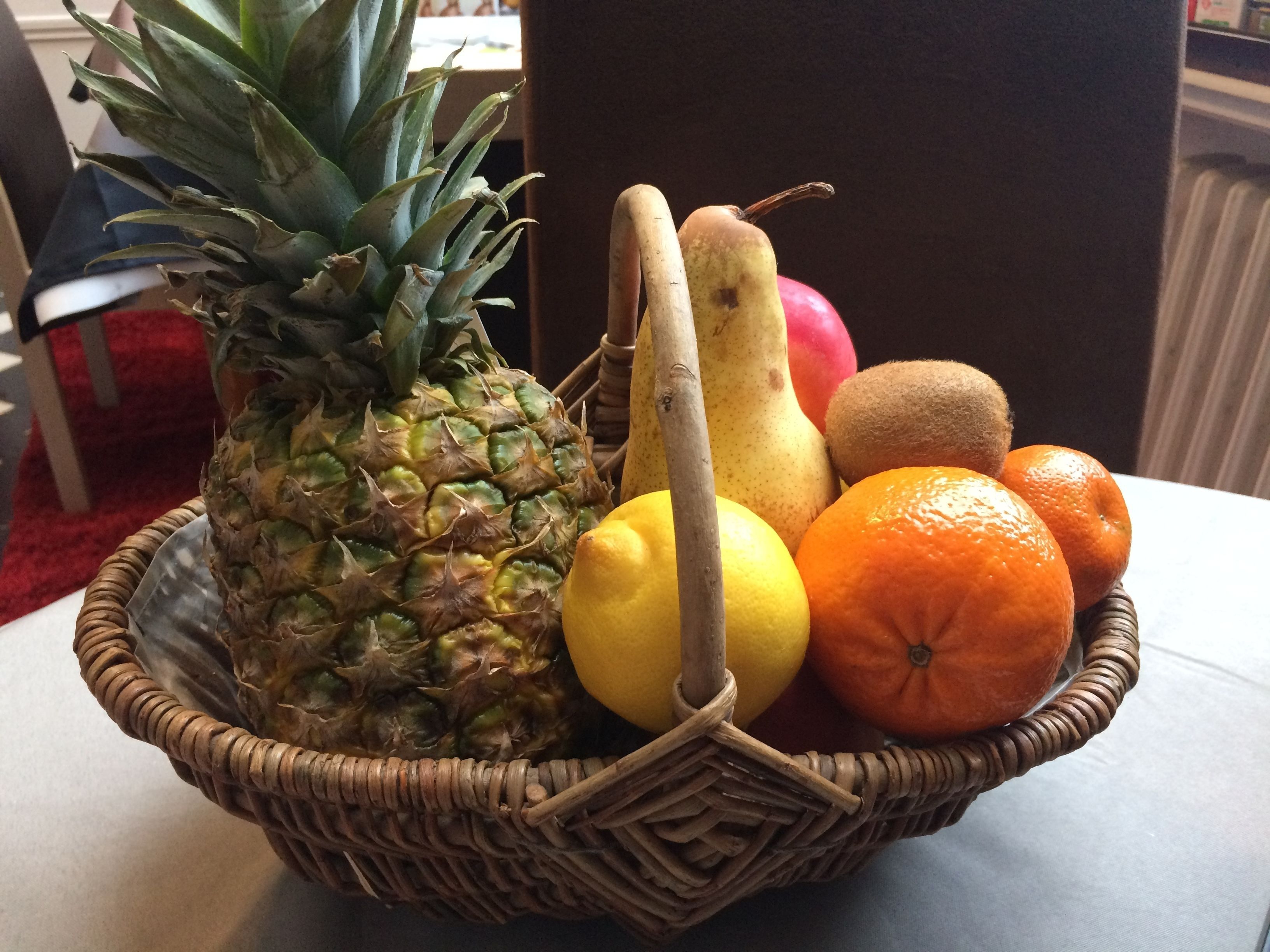 Un panier en osier rempli de fruits, notamment un ananas, un citron, un kiwi, une poire, une pomme et des oranges.