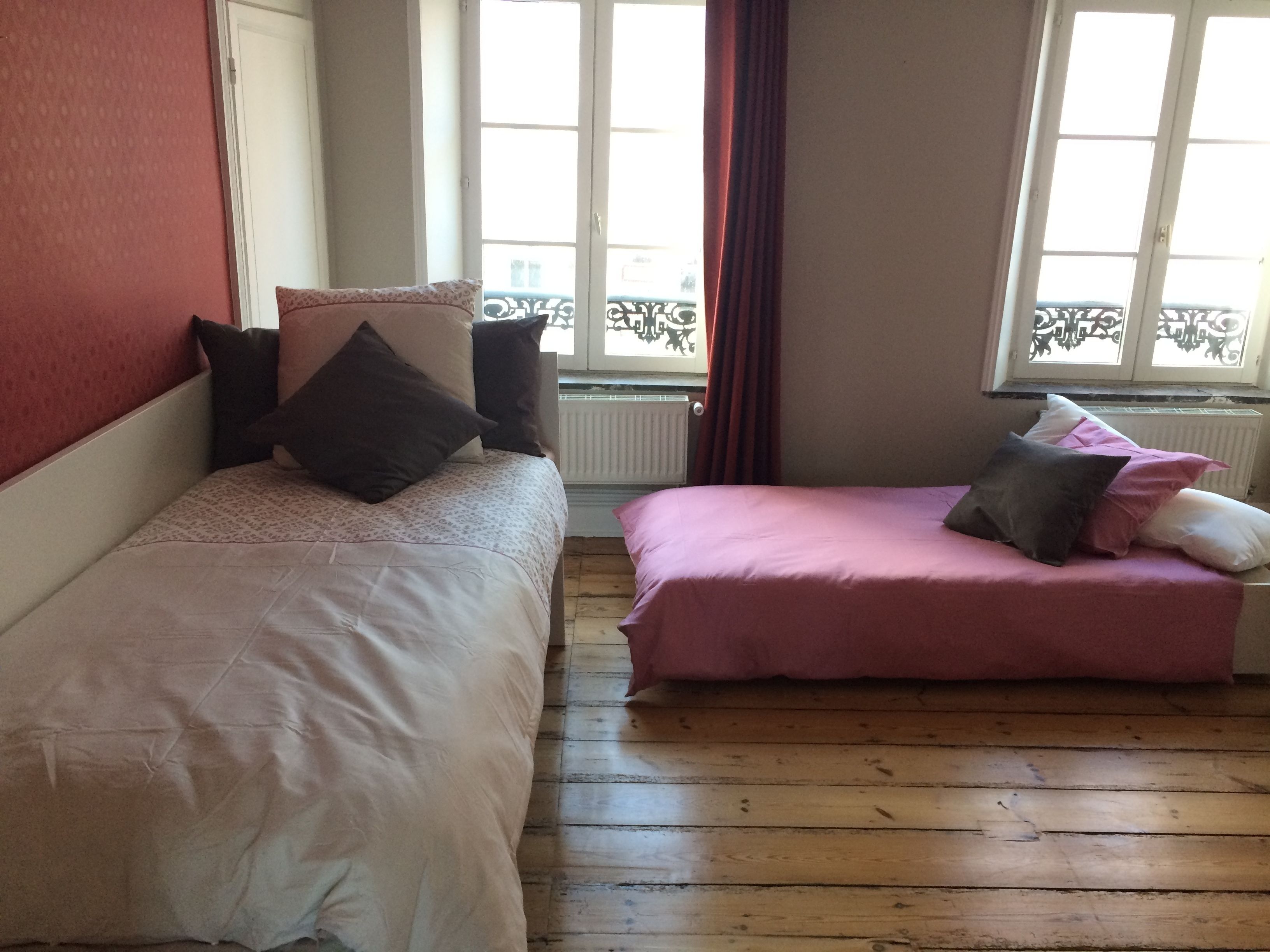 A room with two single beds, each with a patterned bedspread and pillows. The room has wooden flooring and large windows with curtains.