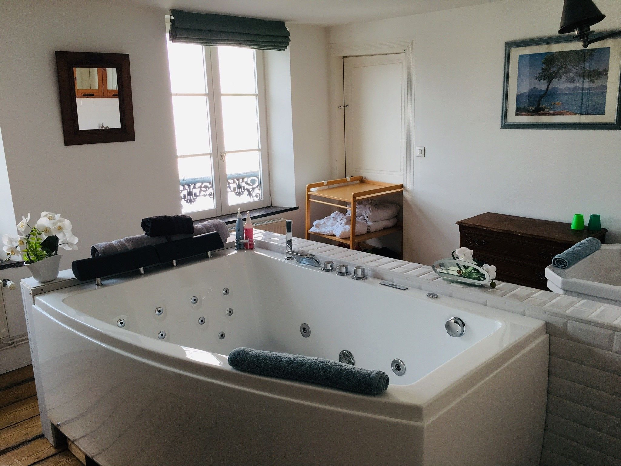 A modern bathroom with a large bathtub, a wooden shelf with towels, and a framed picture on the wall.