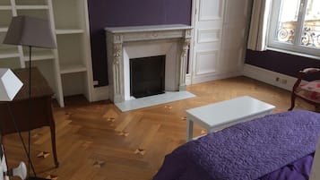 A room with a fireplace, a bed with a purple blanket, a white table, and a chair with a floral cushion.