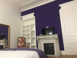 A bedroom with a bed, a dresser, a television on a mantel, and a fireplace.