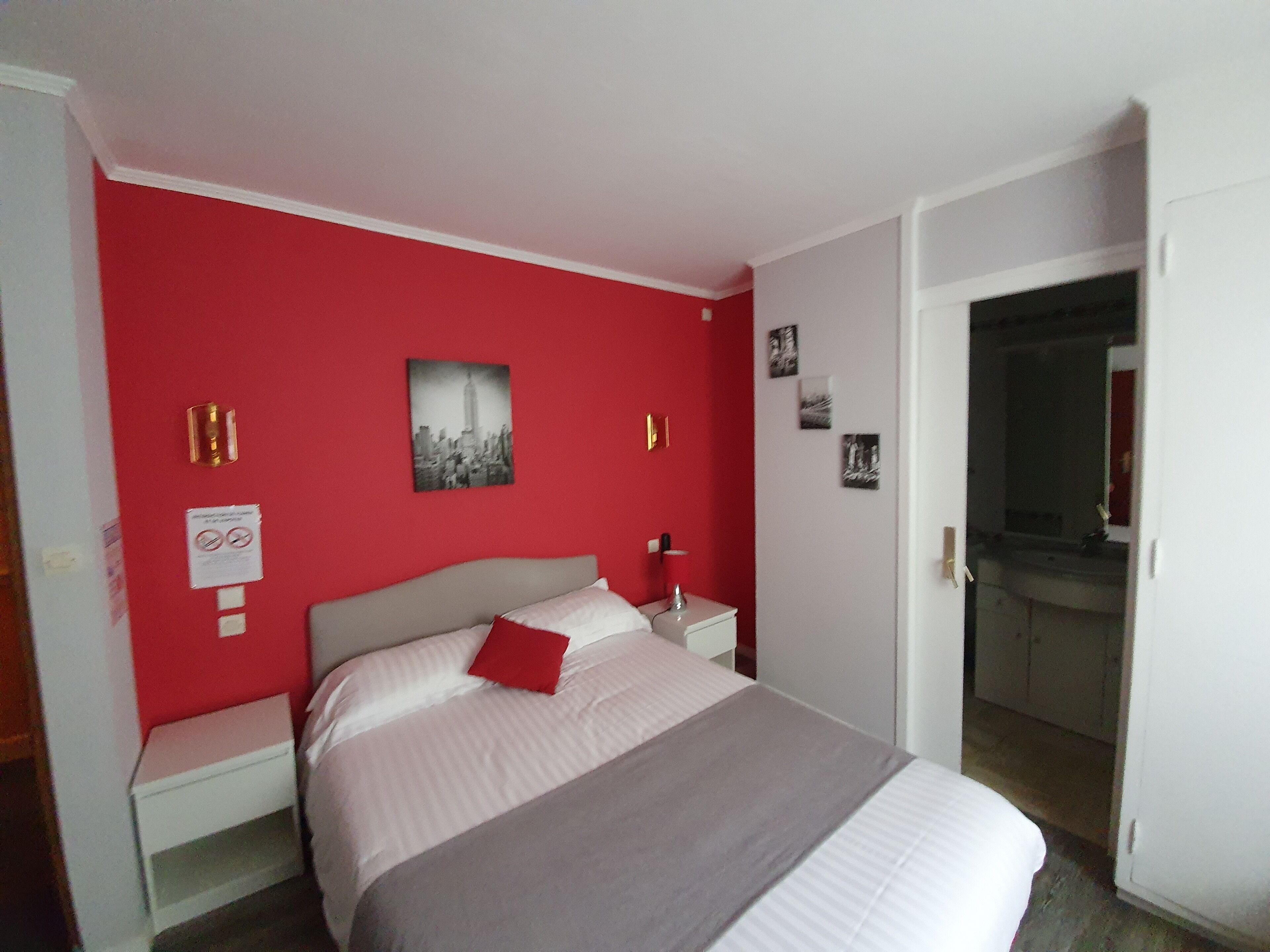 Un dormitorio con una pared roja, una cama con ropa de cama blanca y una almohada roja, una mesita de noche, un cuadro colgado en la pared y una puerta que da a otra habitación.