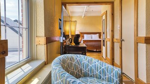 Una habitación de hotel con un ventanal grande, un sillón estampado, una mesita de noche con lámpara y una puerta de madera.