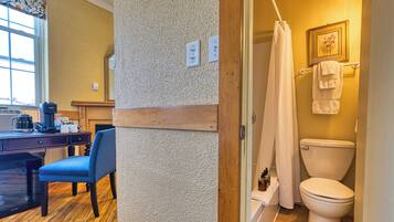 A bathroom with a toilet, shower curtain, and a towel rack. There is a chair and a desk with a coffee maker and a mug.