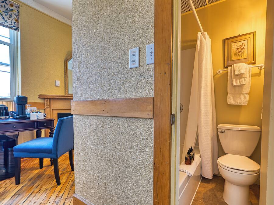A bathroom with a toilet, shower curtain, and a towel rack. There is a chair and a desk with a coffee maker and a mug.