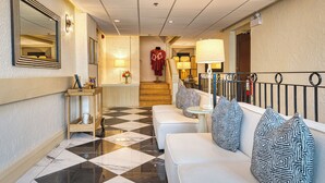 Un vestíbulo de hotel con suelo a cuadros, una recepción, una escalera y una zona de estar con cojines estampados.