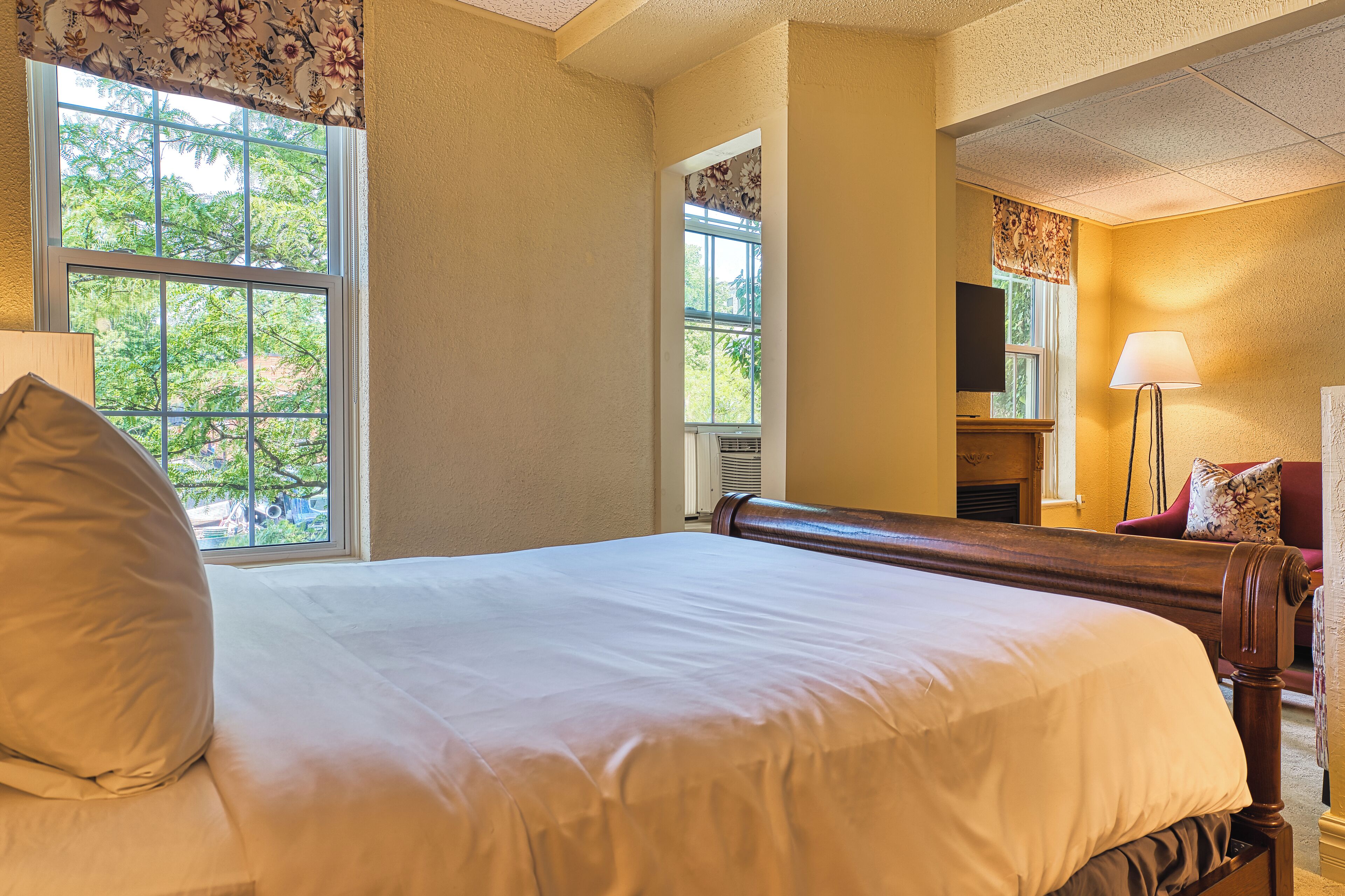 Une chambre d’hôtel avec un grand lit, une chaise, une lampe et une fenêtre donnant sur des arbres.