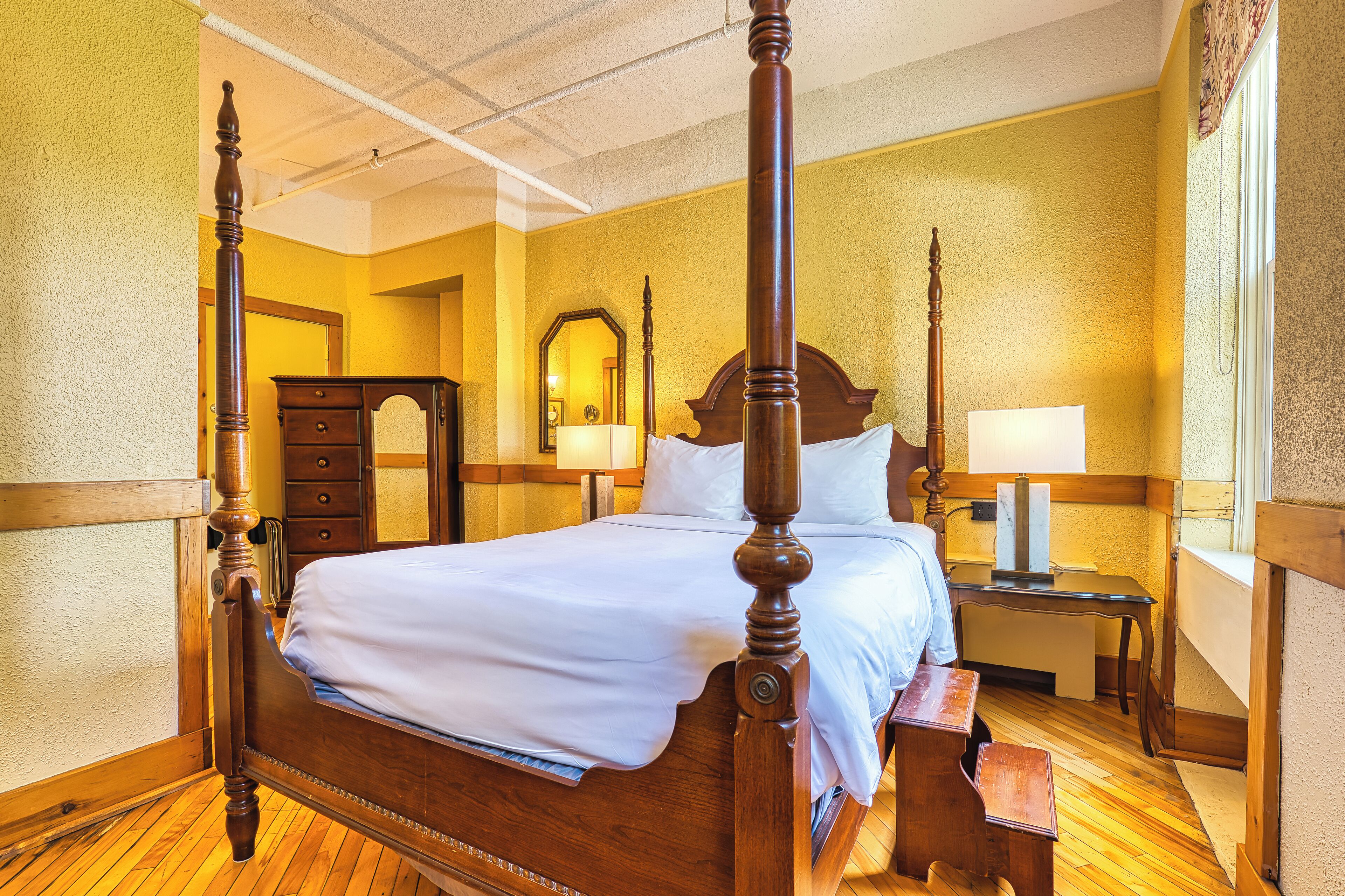 A four-poster bed with a wooden headboard, a nightstand with a lamp, and a wooden dresser in a room with yellow walls and wooden flooring.