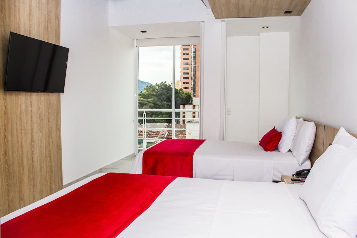 Ein Hotelzimmer mit zwei Betten, einem an der Wand montierten Fernseher und einem Balkon mit Blick auf Gebäude und Bäume.