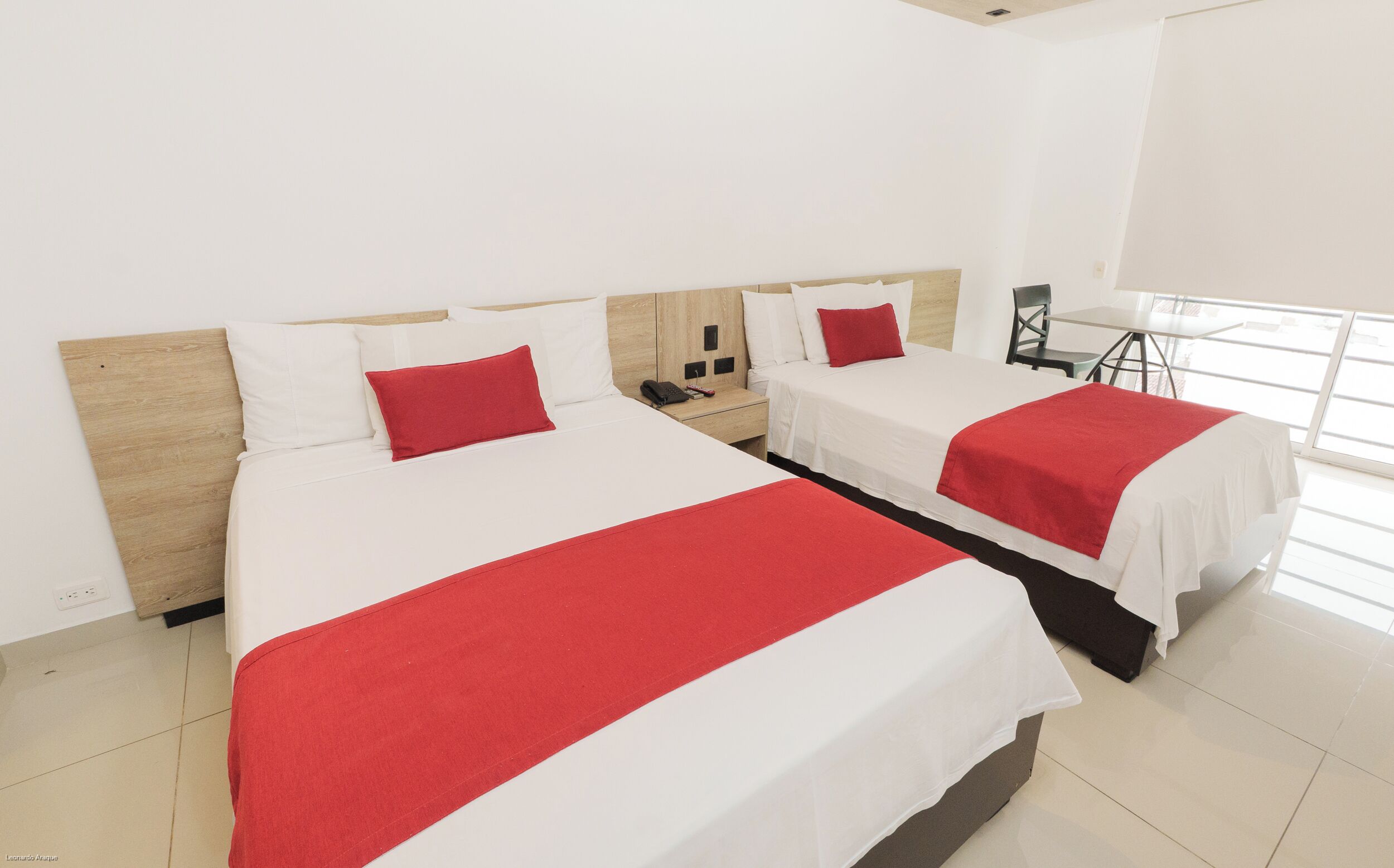 Een hotelkamer met twee bedden, elk voorzien van witte beddengoed en rode accenten. In de hoek staat een klein tafeltje en een stoel, en een raam zorgt voor natuurlijk licht.