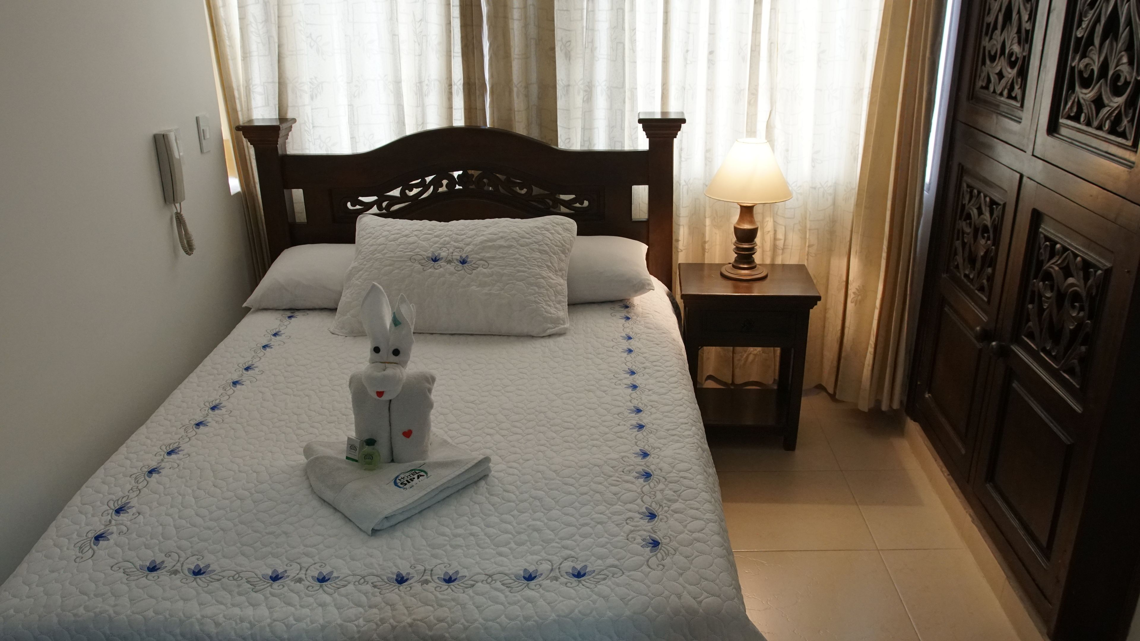 A neatly made bed with a white quilt, a decorative pillow, and a towel shaped like a rabbit. There is a bedside lamp on a wooden nightstand.