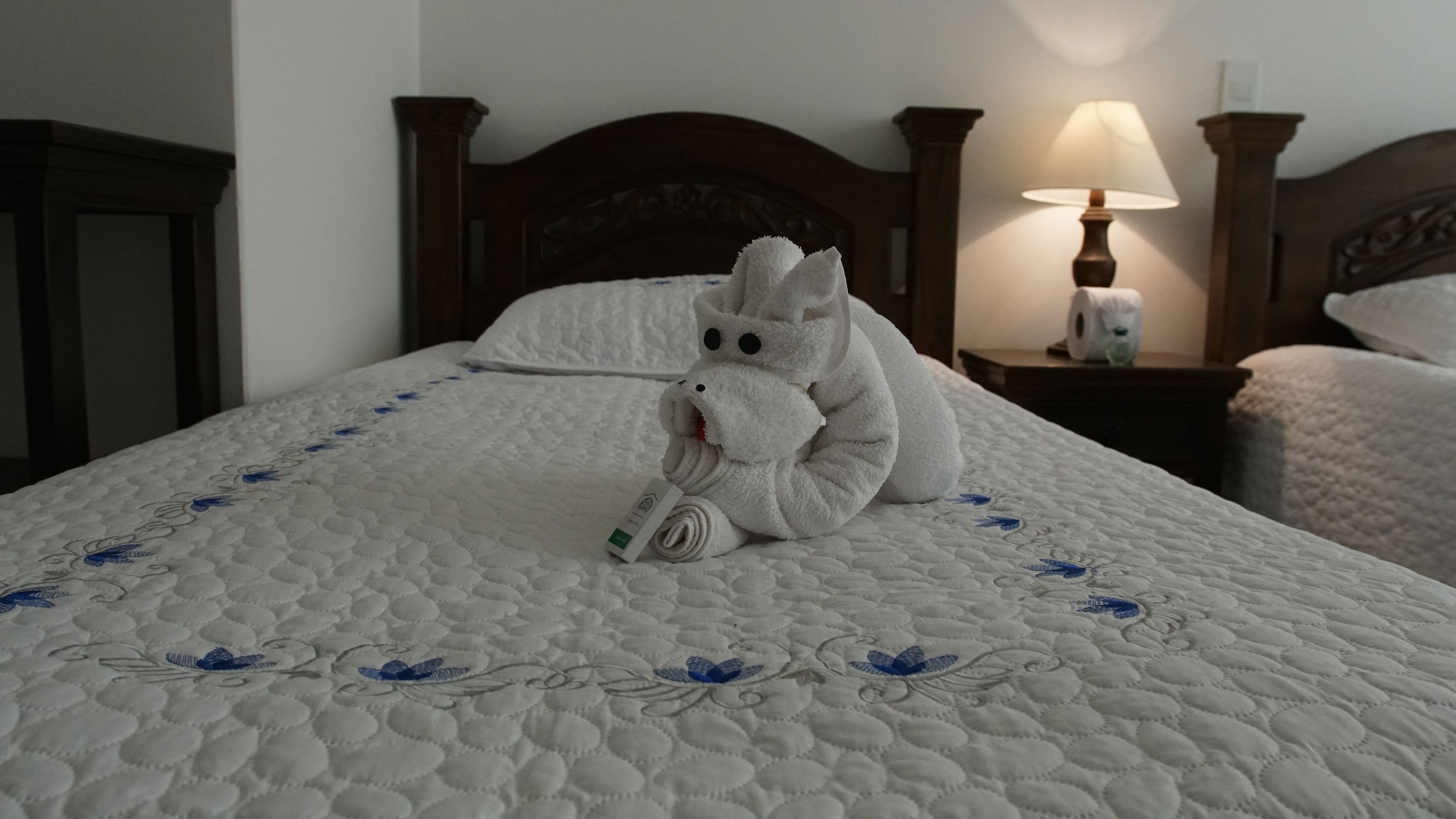 A neatly made bed with a white quilt and blue floral patterns, a wooden headboard, a bedside lamp, and a rolled-up towel shaped like a bear.