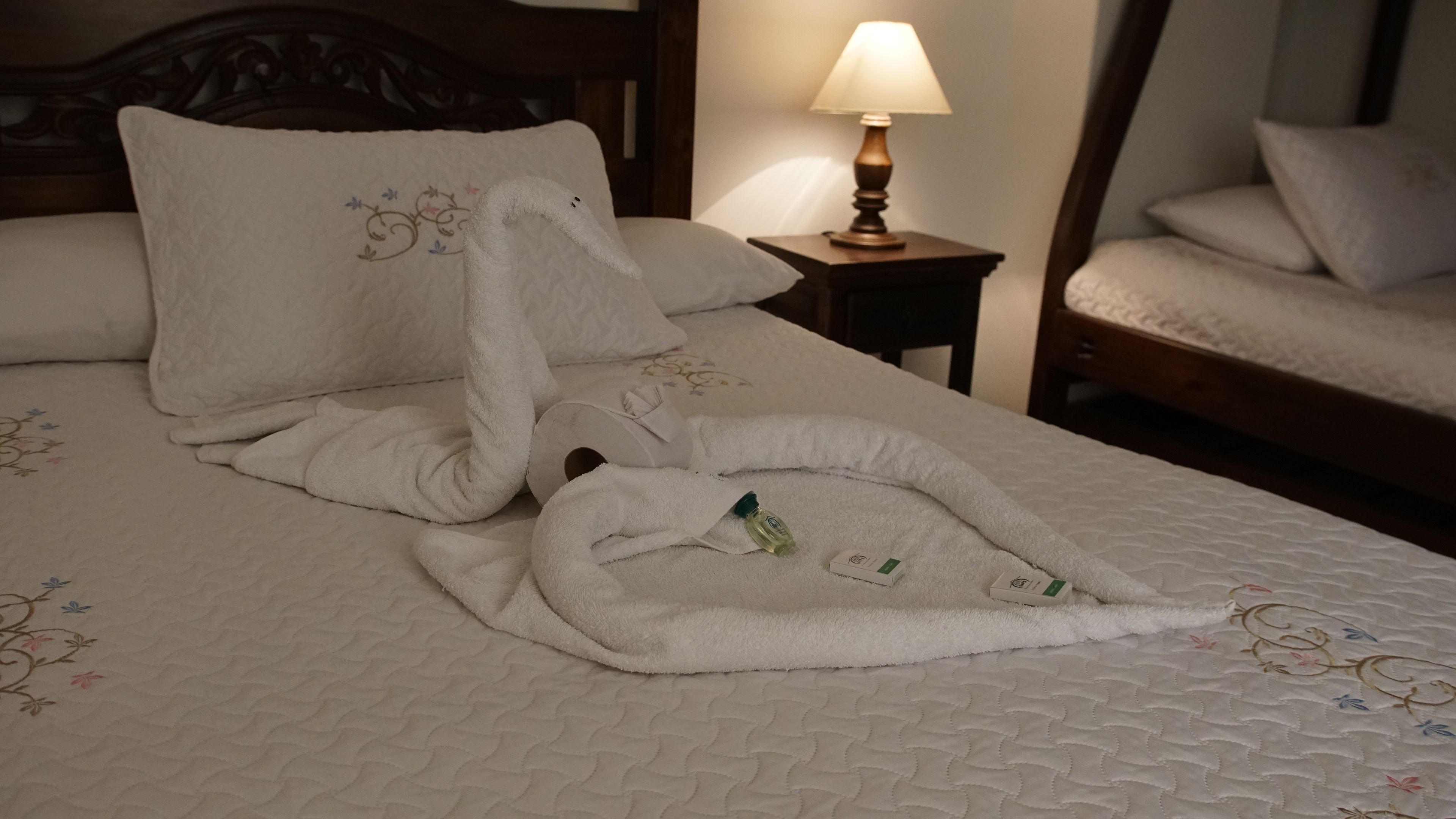 Una cama bien hecha con edredón blanco, una botella de loción y una pequeña caja, probablemente con cepillo e pasta de dientes.