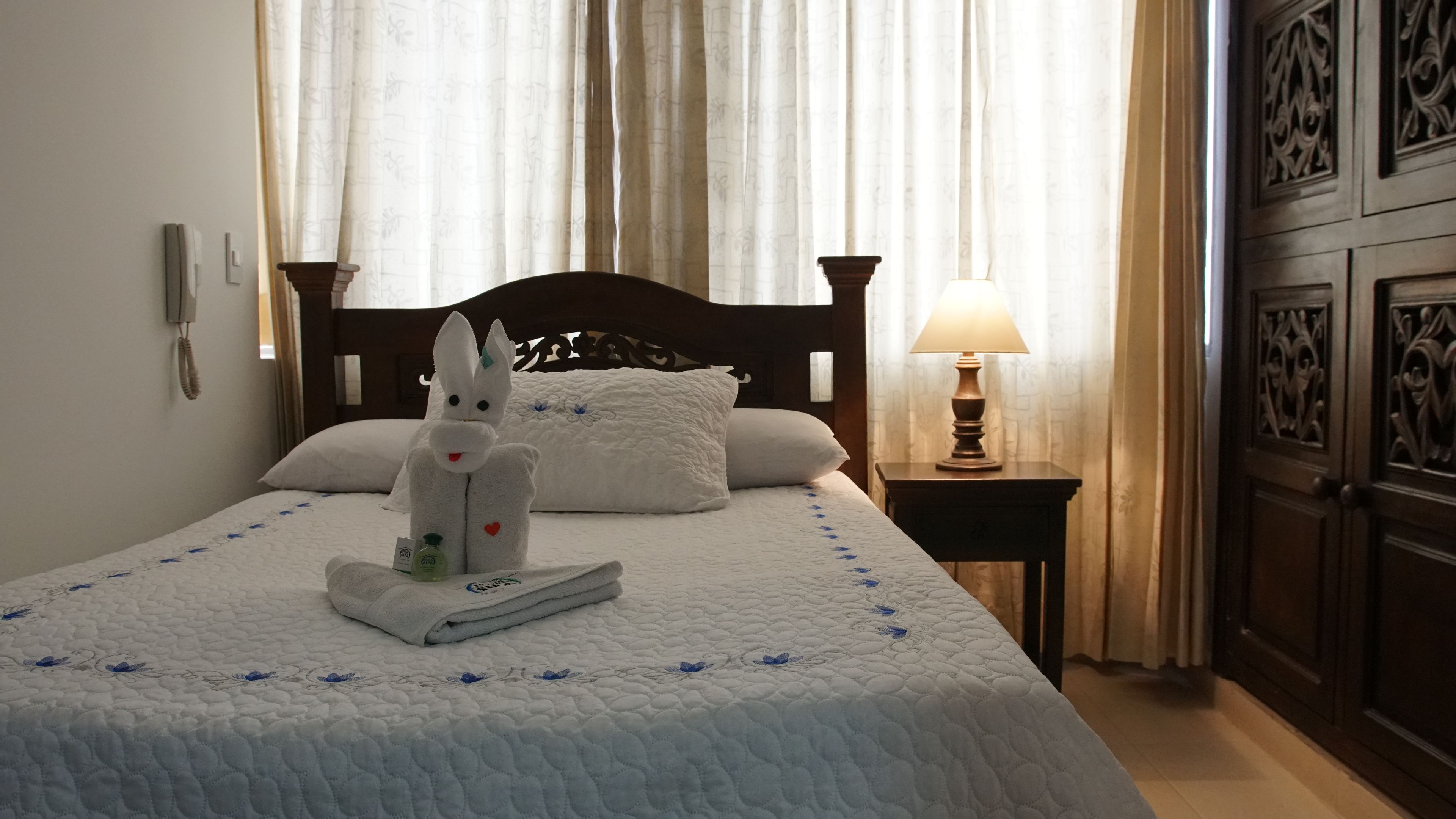 A hotel room with a bed, a nightstand with a lamp, a towel animal, and a bottle of lotion.
