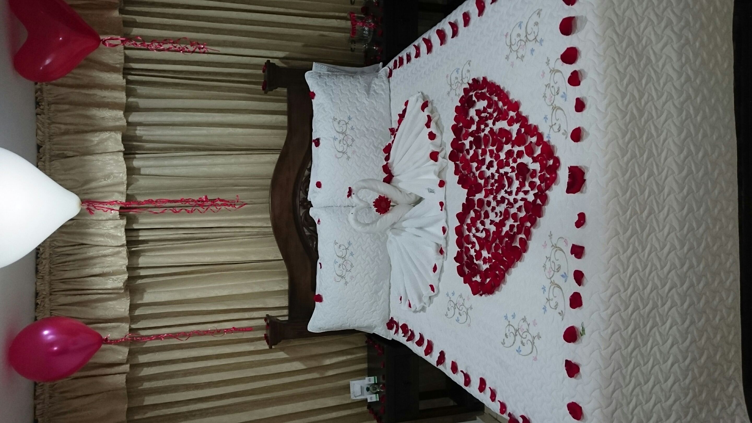 Una habitación con una cama cubierta con un colcha blanca, una decoración con forma de corazón hecha de pétalos de rosa roja y globos rojos.
