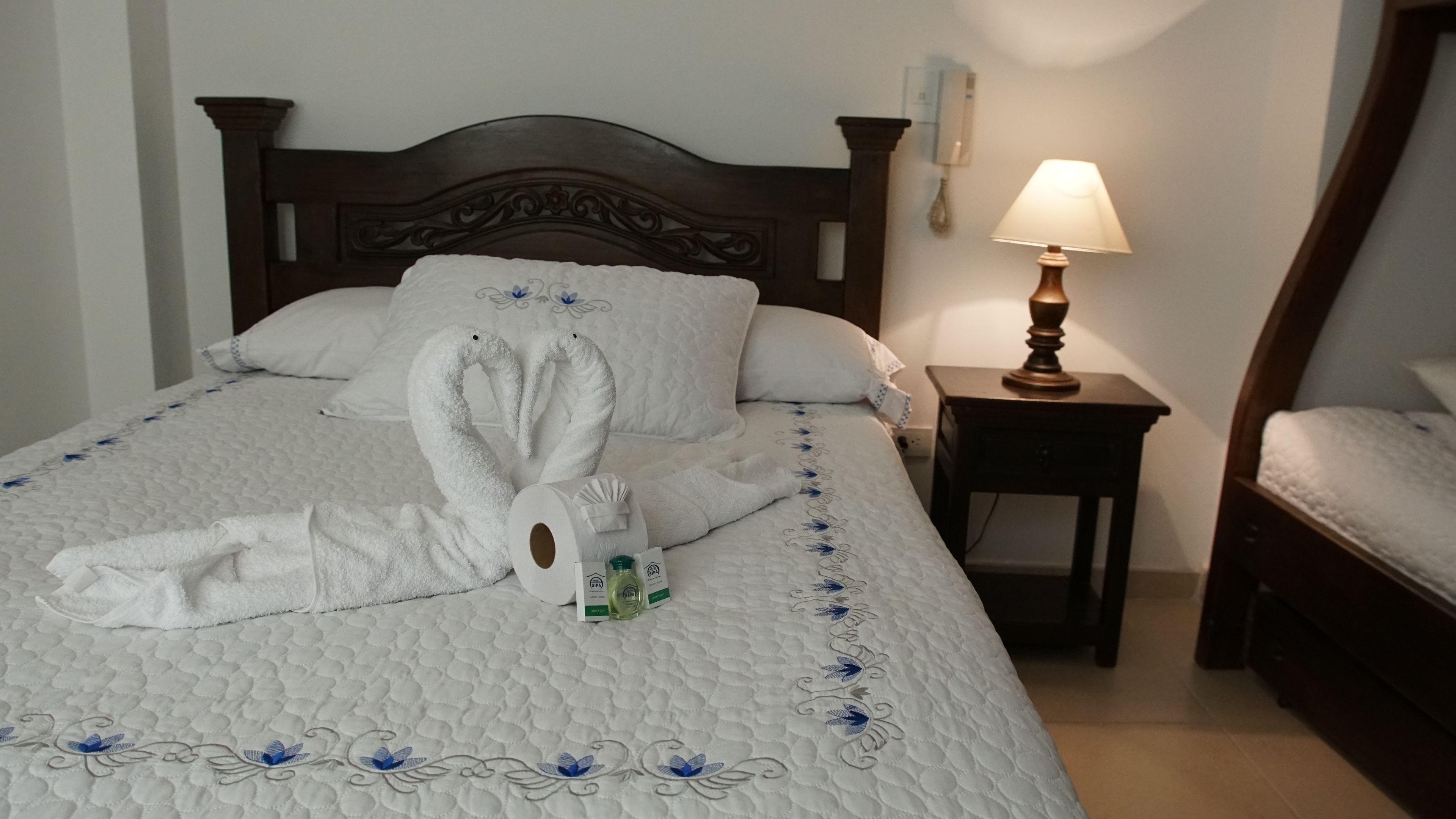 A neatly made bed with a white duvet and pillows, a wooden headboard, a bedside table with a lamp, and a small nightstand.