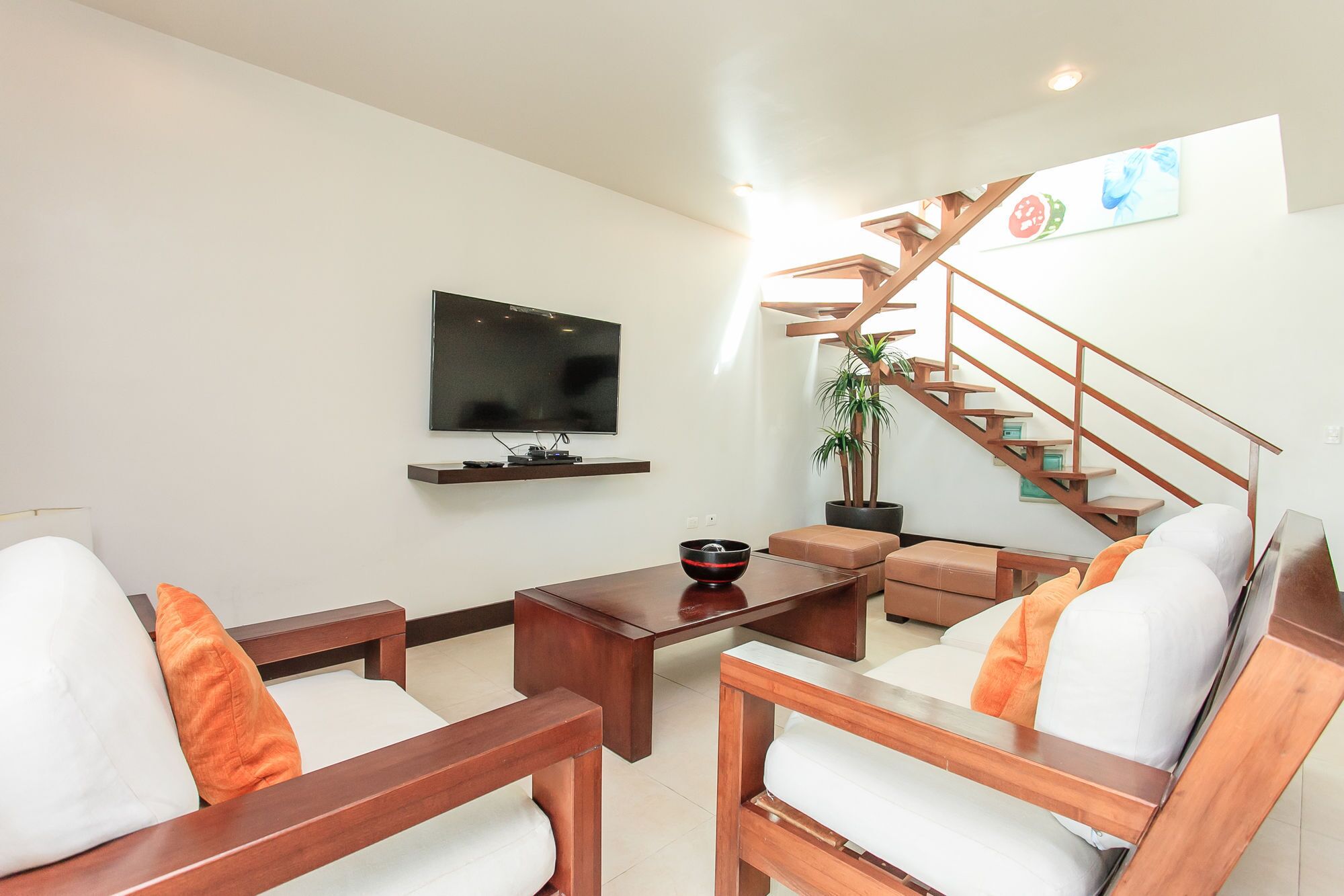 A modern living room with a flat-screen TV, wooden furniture, a staircase, and a potted plant.