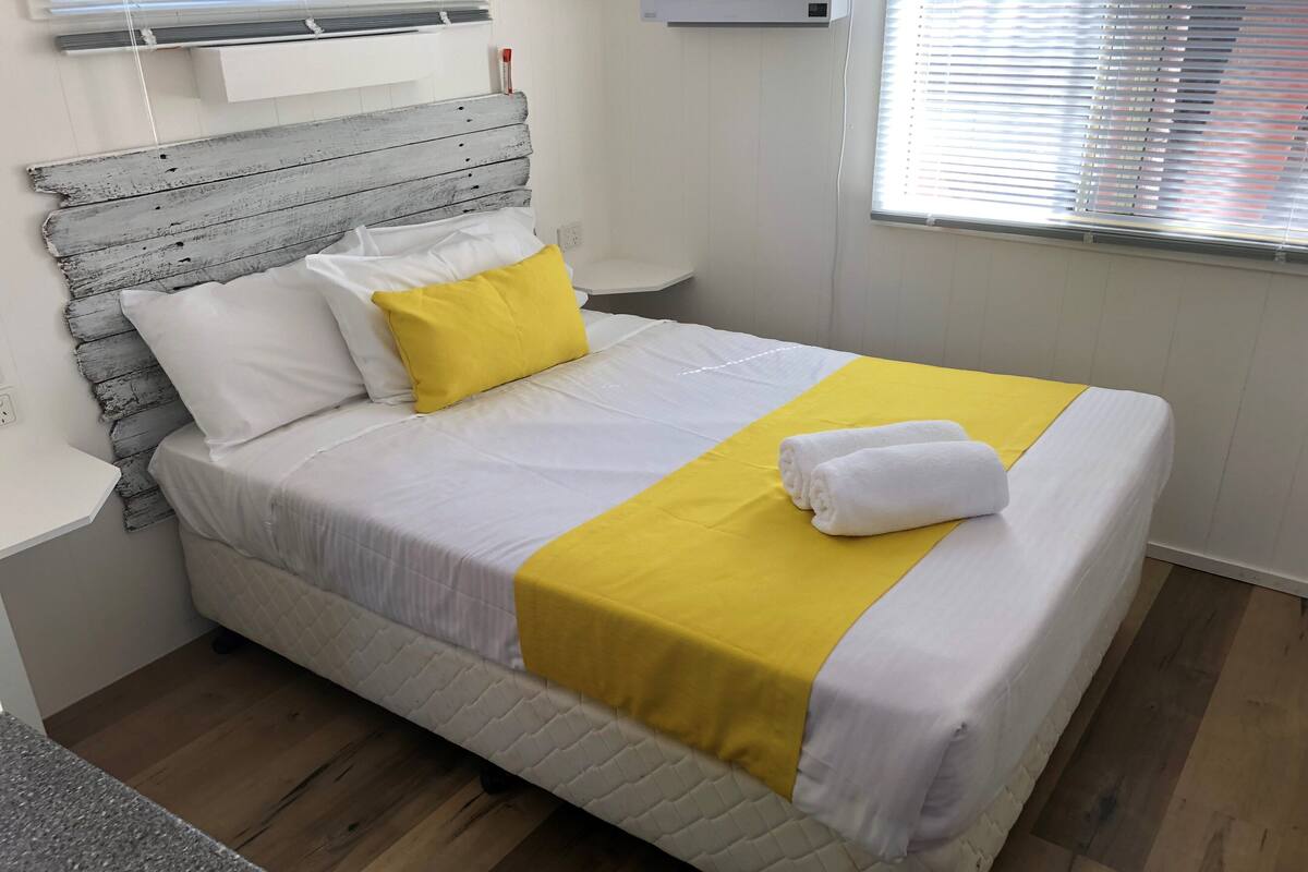 A neatly made bed with white and yellow bedding, a wooden headboard, and a window with blinds.