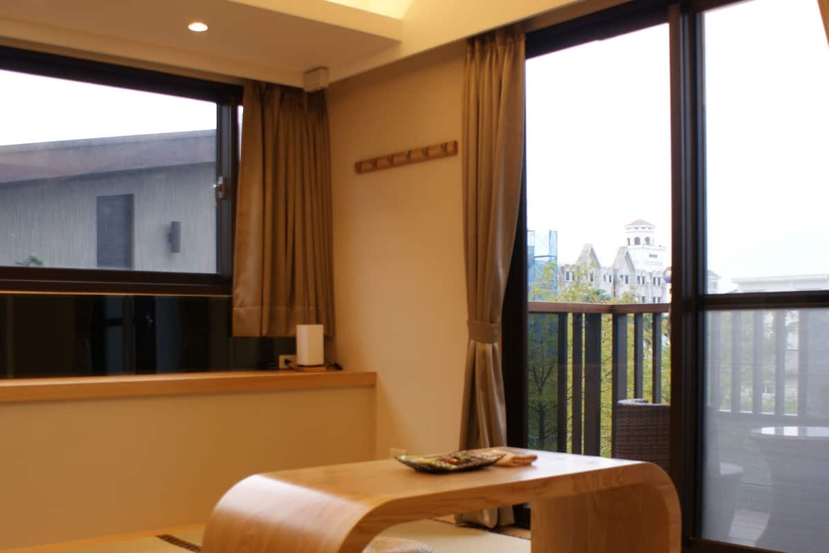 Una habitación con esteras de tatami, una mesa de madera baja, un taburete tejido y una lámpara colgante.