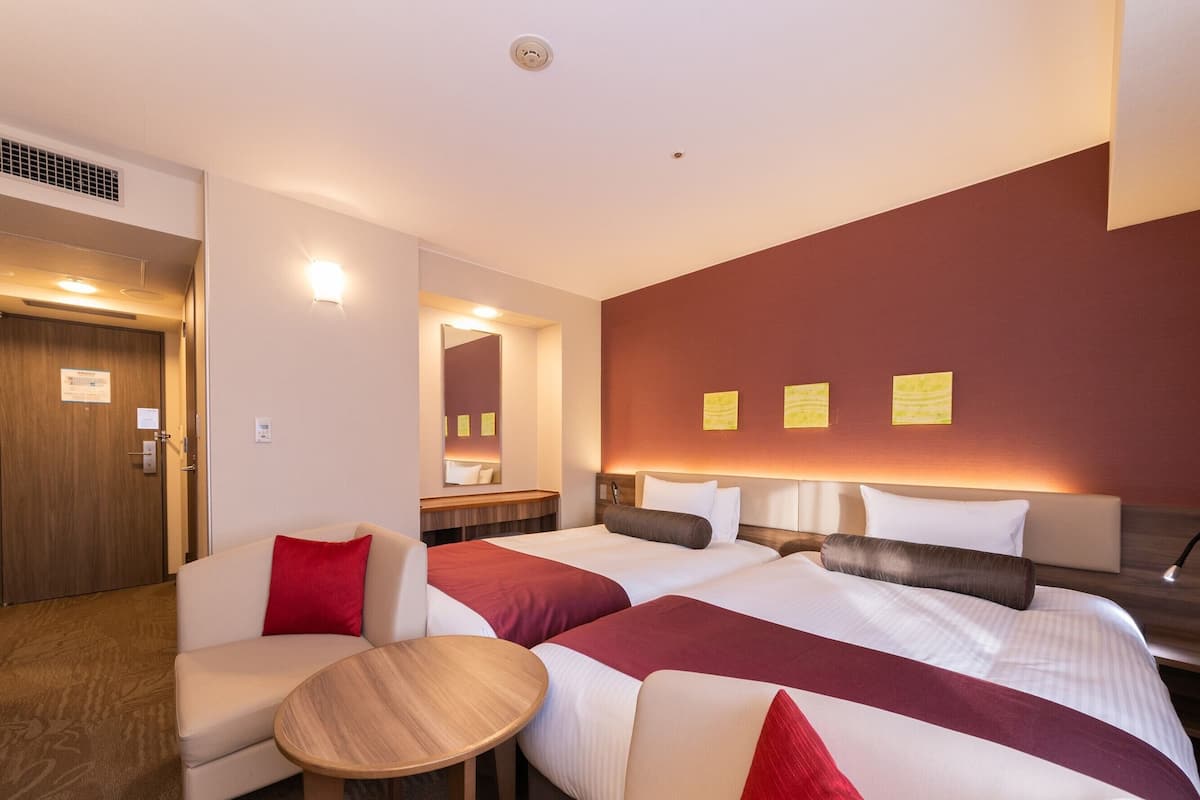A hotel room with two beds, a sitting area with a table, a mirror, and a red accent wall.