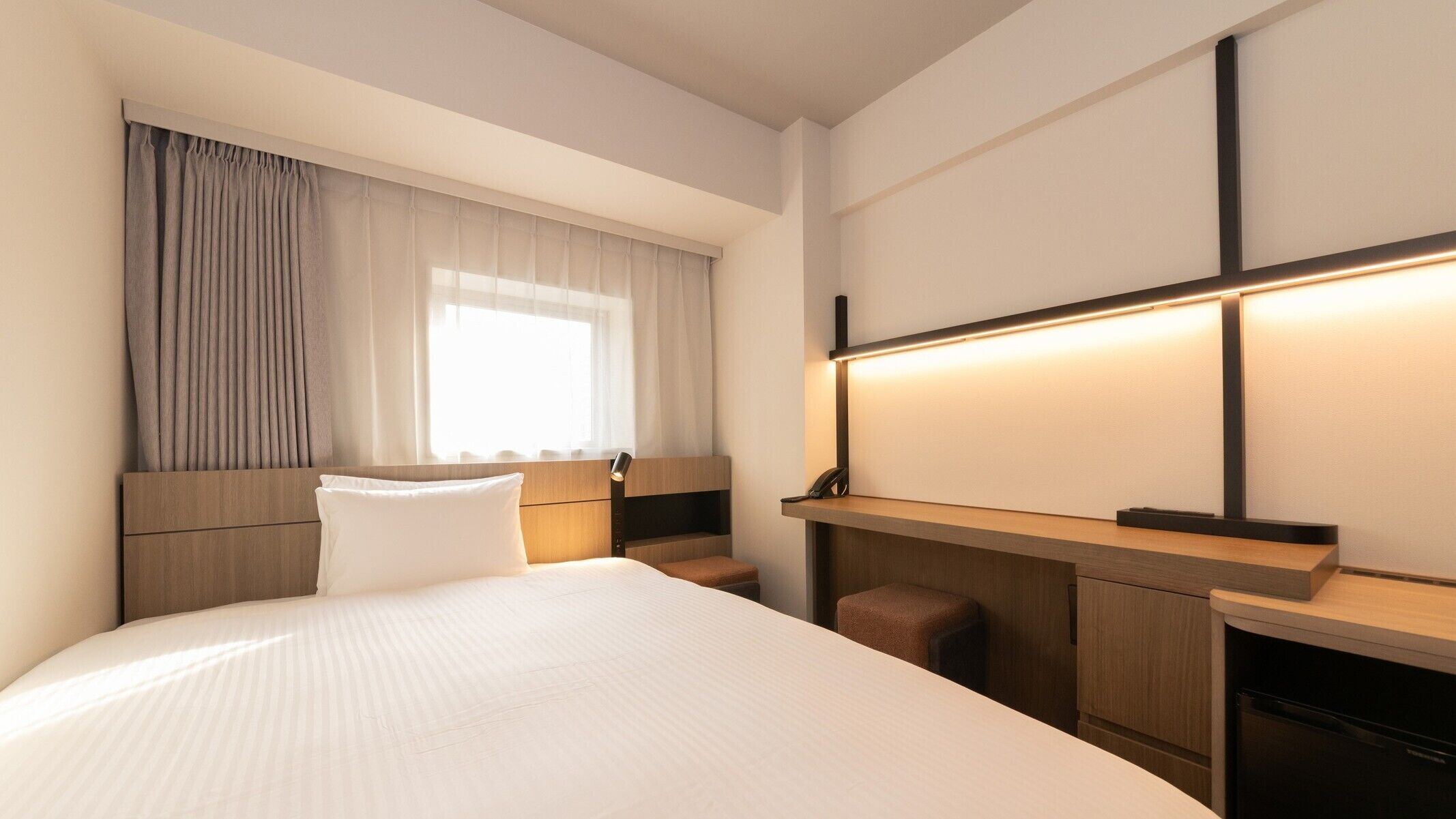 A hotel room with a large bed, a bedside table, a desk, a chair, a window with curtains, and a wall-mounted light.