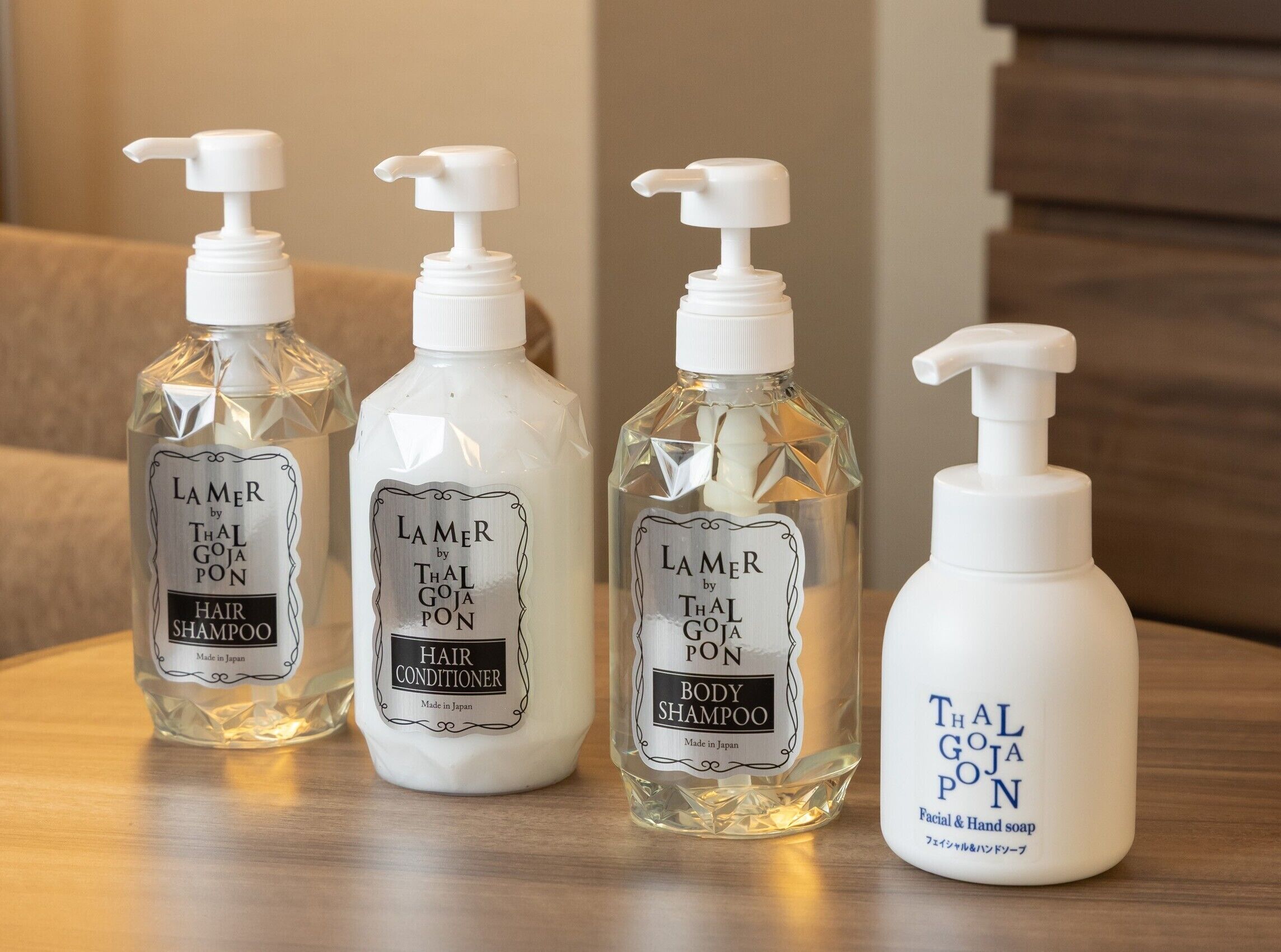 Cuatro botellas de productos Lamer by Thal Goja: champú para cabello, acondicionador para cabello, champú corporal y jabón facial y para manos, dispuestas sobre una superficie de madera.