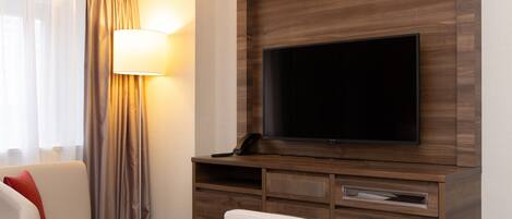 A hotel room with a flat-screen TV mounted on a wooden wall, a bedside table, a lamp, and a sofa.