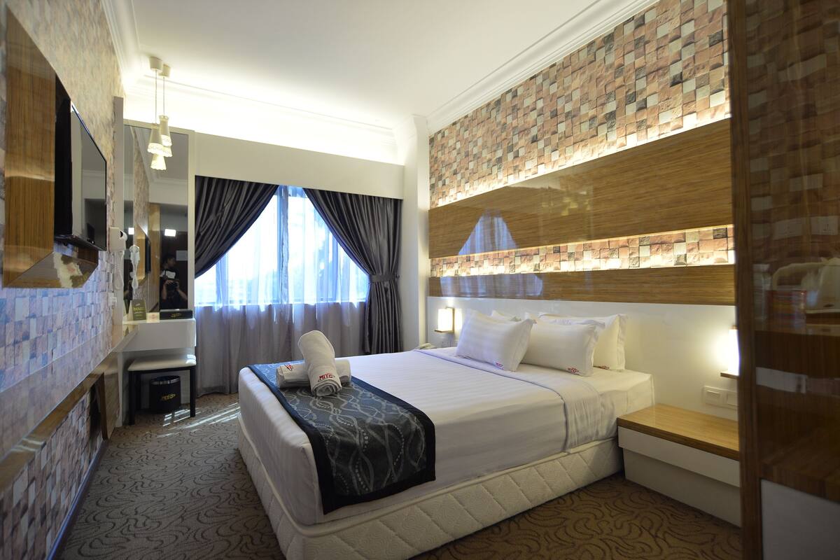 A hotel room with a large bed, a bedside table, a mirror, a window with curtains, and a wall with a mosaic pattern.