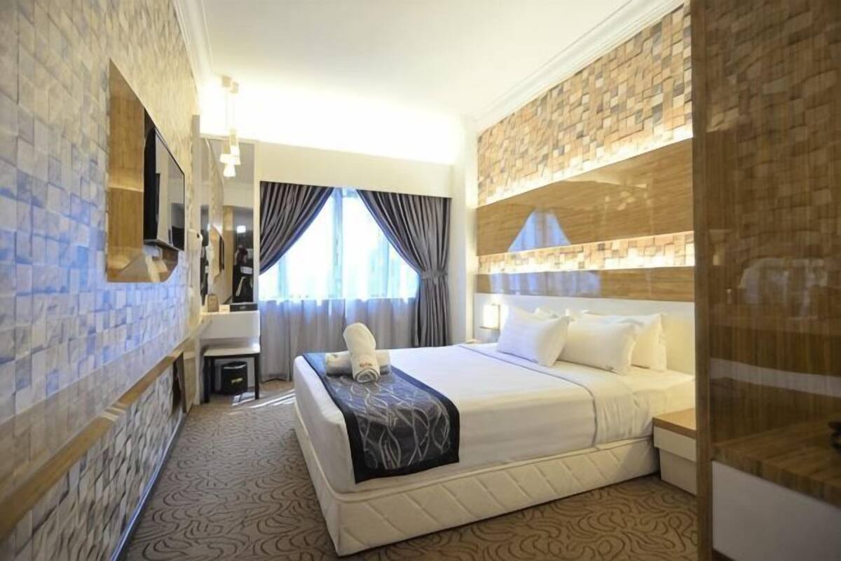 A hotel room with a bed, a mirror, a small table, a nightstand, a window with curtains, and a wall with a stone-like pattern.