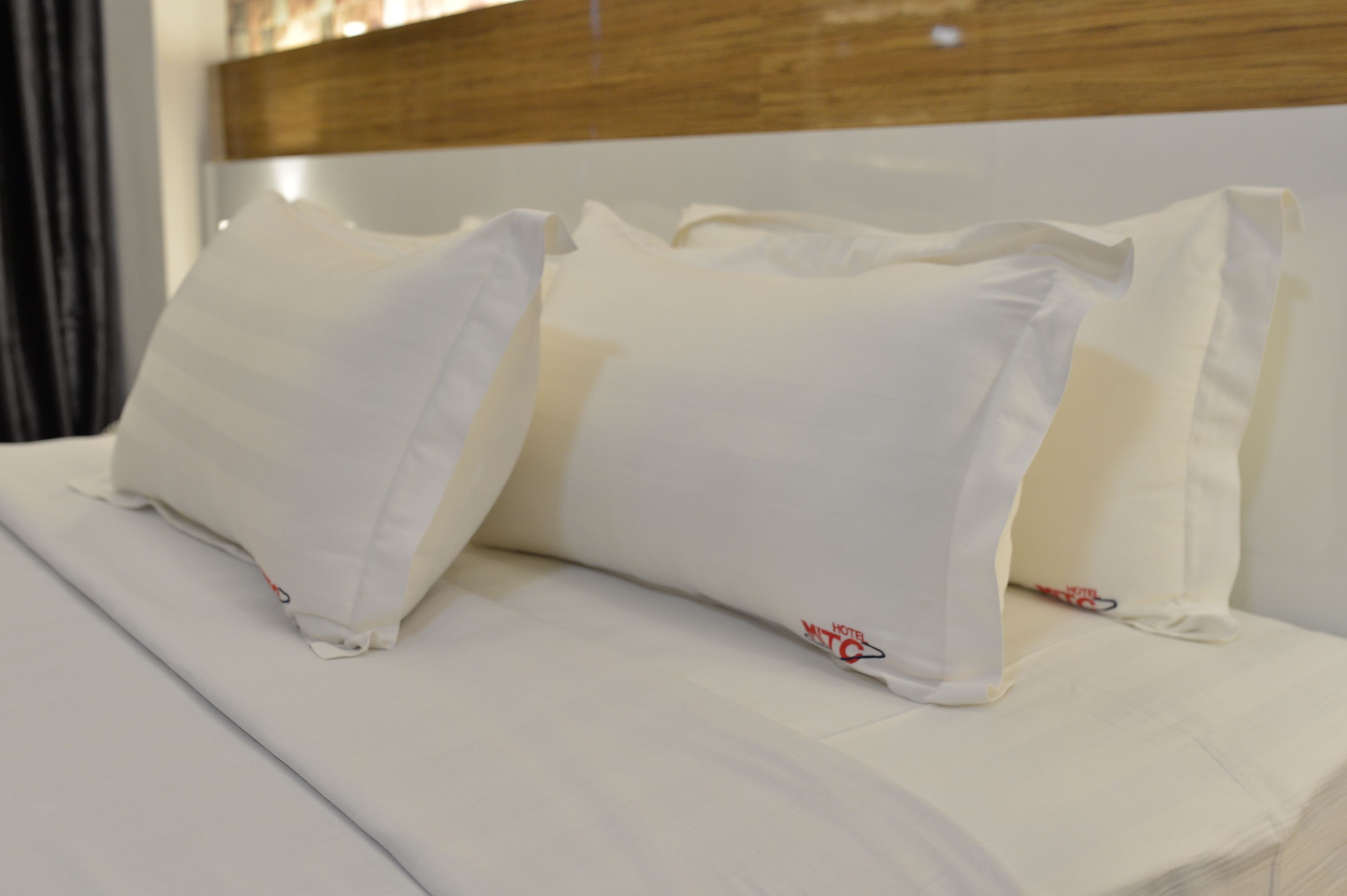 Una cama bien tendida con sábanas y almohadas blancas, cabecera de madera y logotipo visible en la almohada.