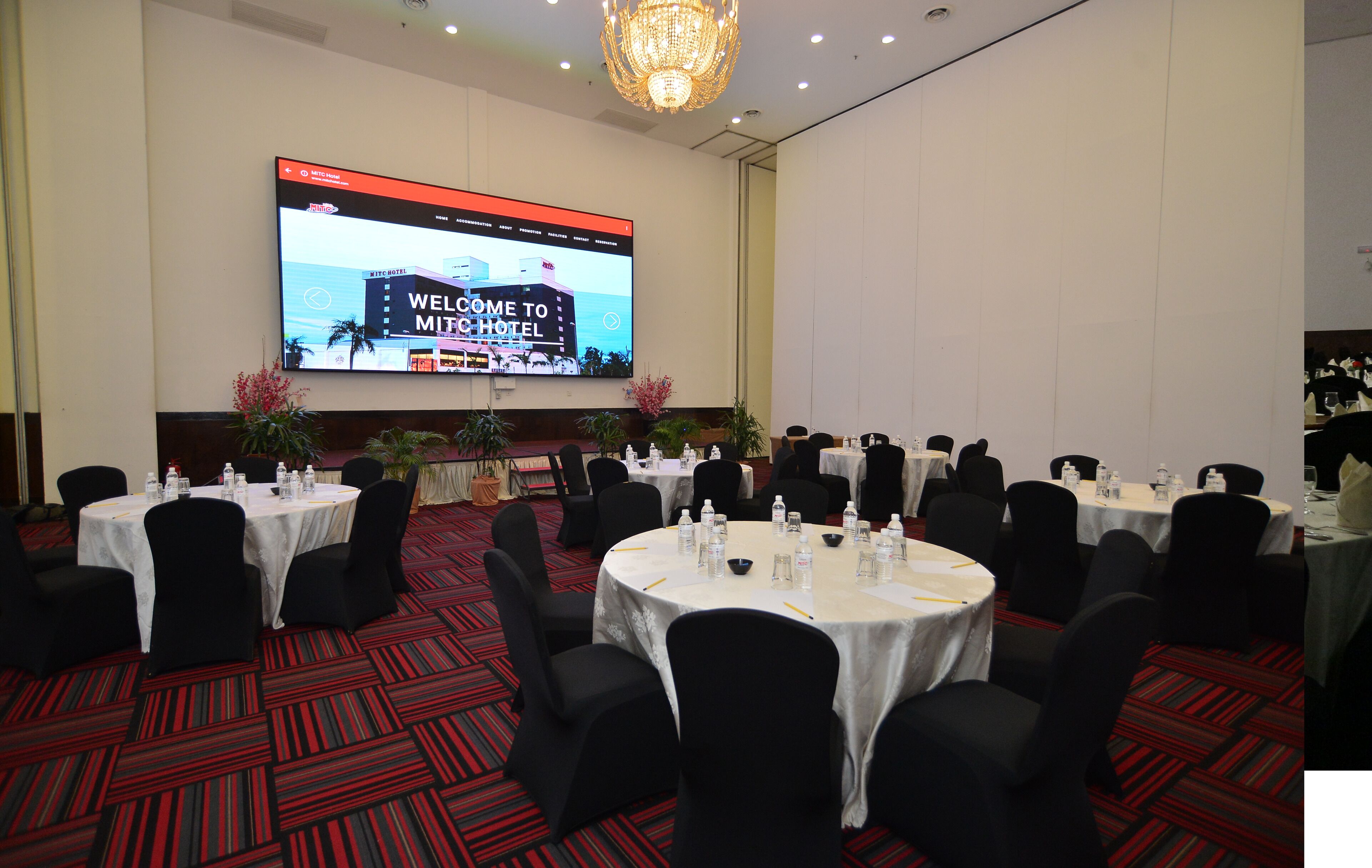 A conference room with multiple round tables, black chairs, and a large screen displaying a welcome message.